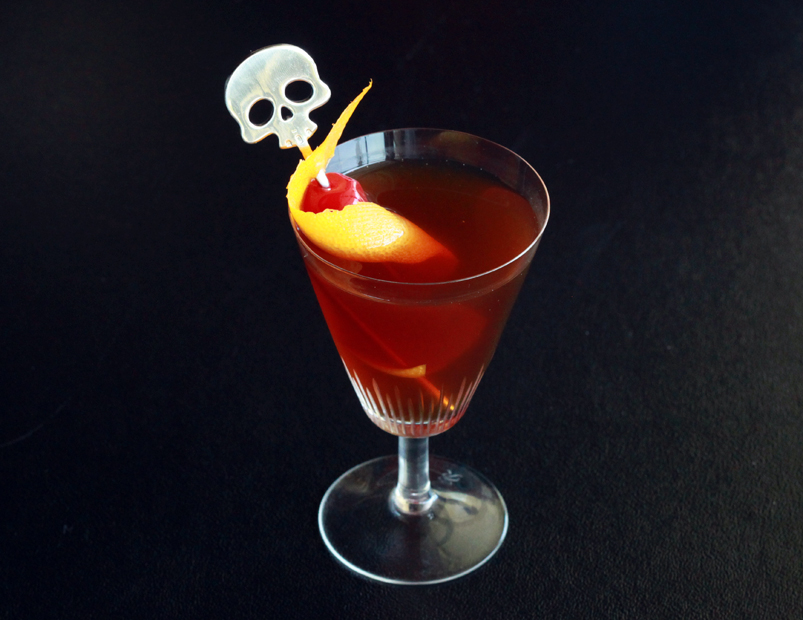 Rise and grind cocktail in a stemmed glass garnished with a maraschino cherry, orange peel, and a skull swizzle stick.