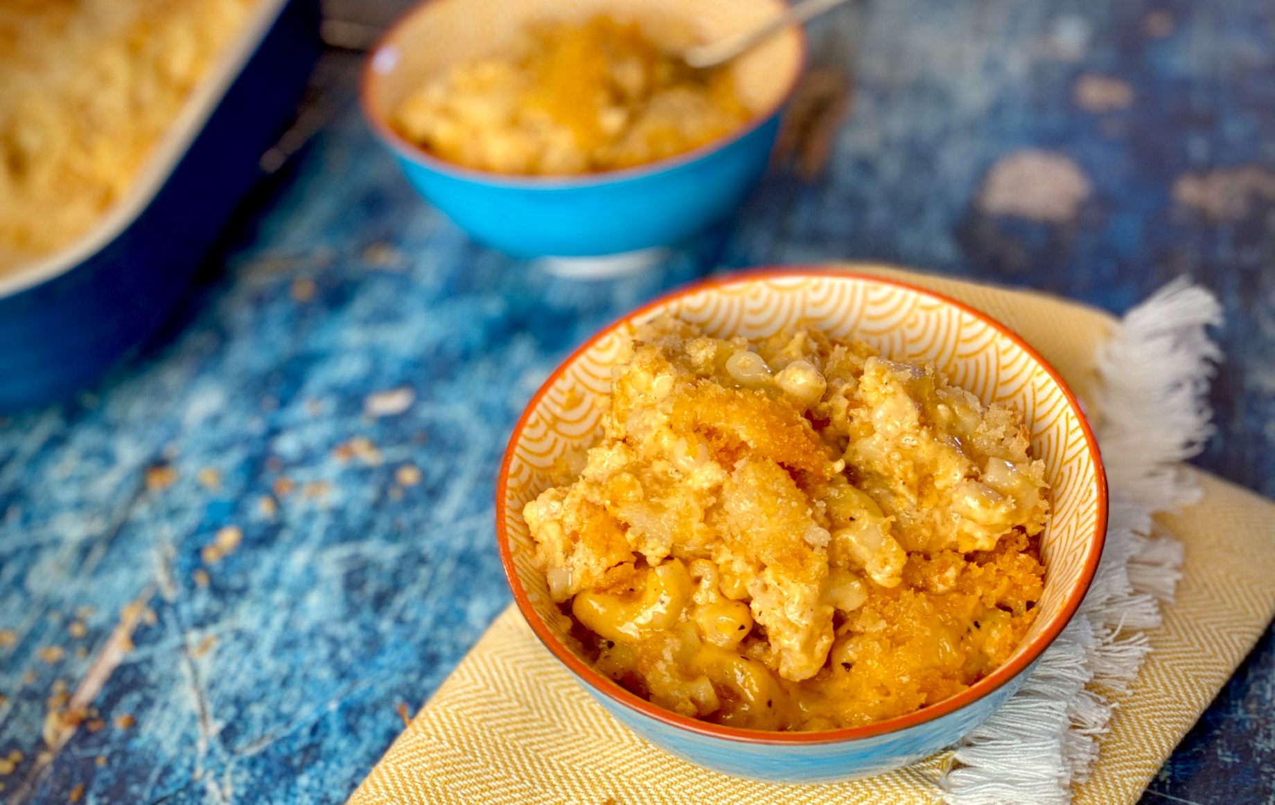 Baked macaroni with bread crumbs in a bowl on a yellow napkin sitting on a bright blue table.