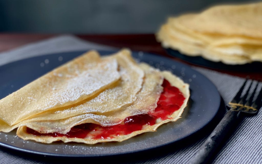 Stack of freshly made crepes filled with strawberry jam served on a blue plate, perfect for brunch or dessert