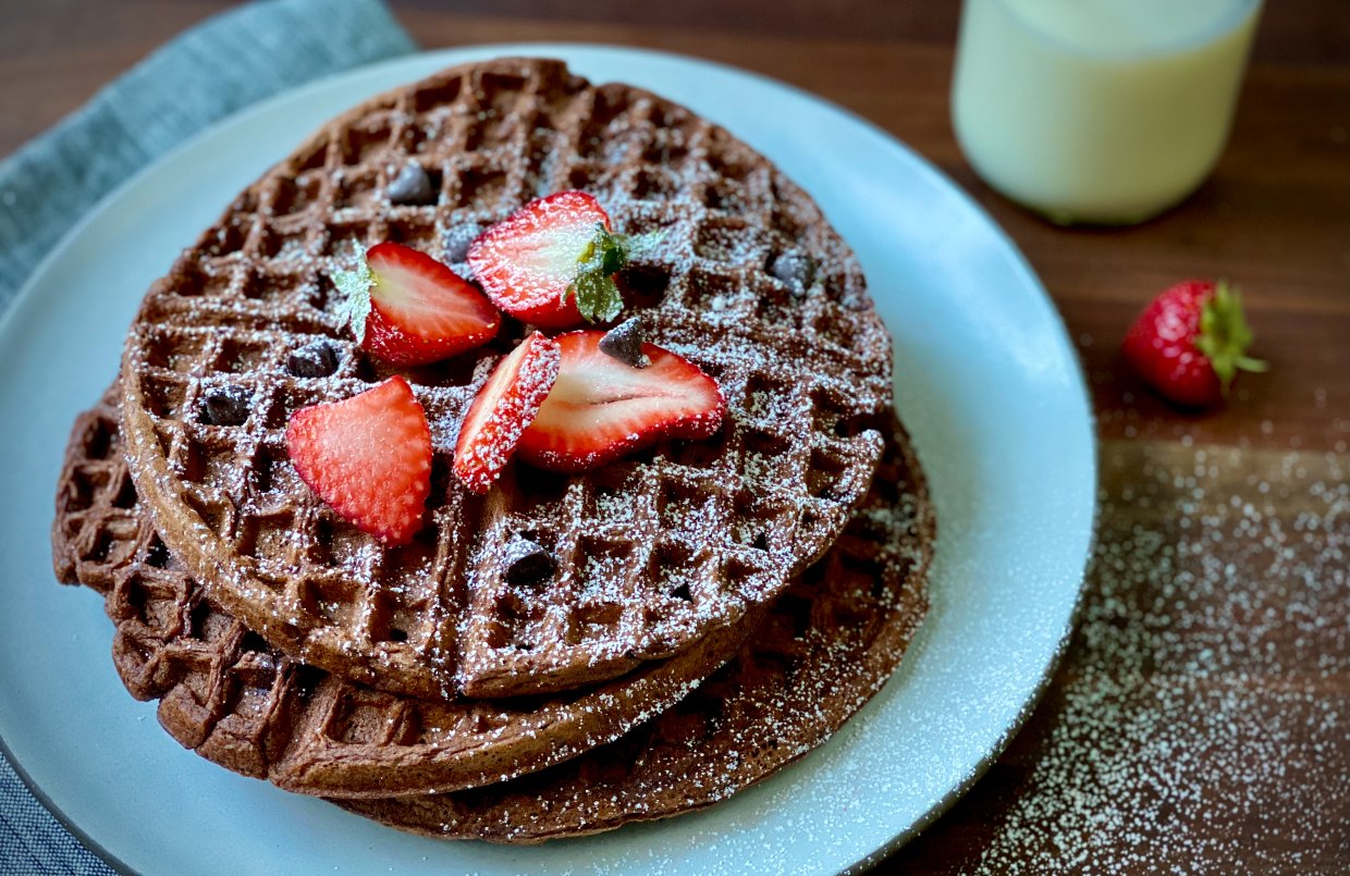 Chocolate waffles stacked on a plate, topped with strawberries and powdered sugar.