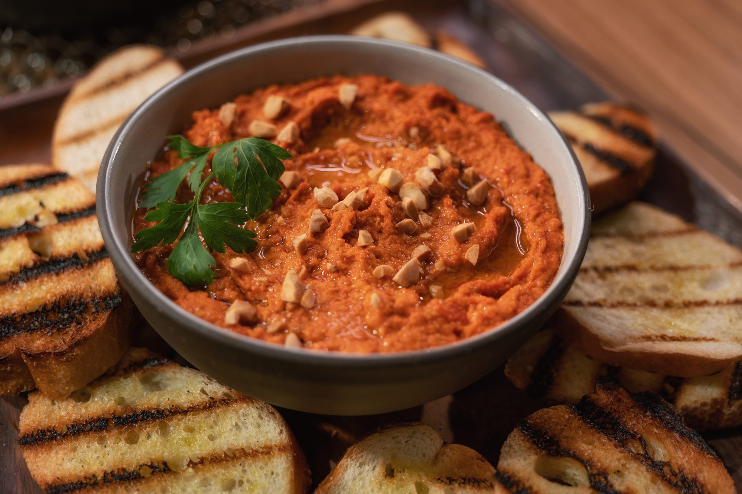 Smoky romesco sauce in a grey bowl surrounded by pieces of grilled, toasted baguette.