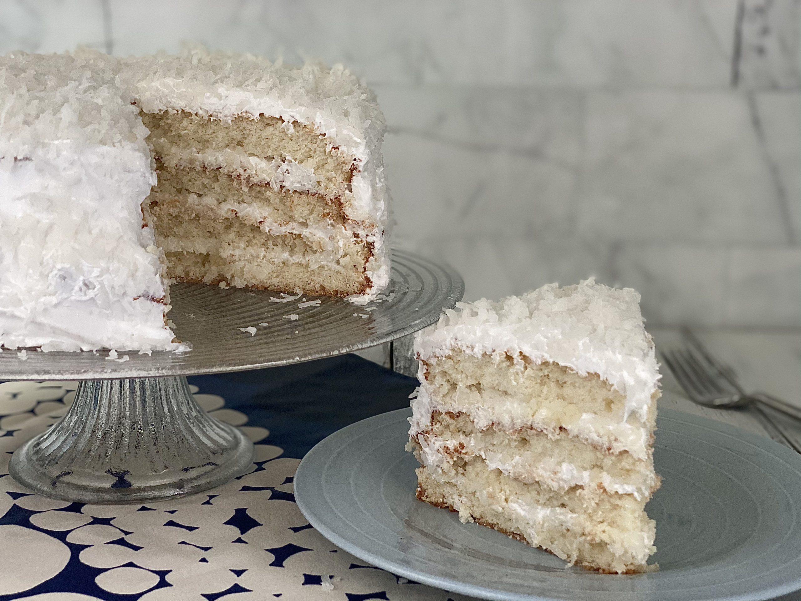 Close-up photograph of a slice of coconut cake with 7-minute frosting. The cake appears moist and fluffy with visible shreds of coconut throughout. The frosting covers the top and sides of the cake, appearing light and fluffy, with toasted coconut flakes sprinkled generously on top