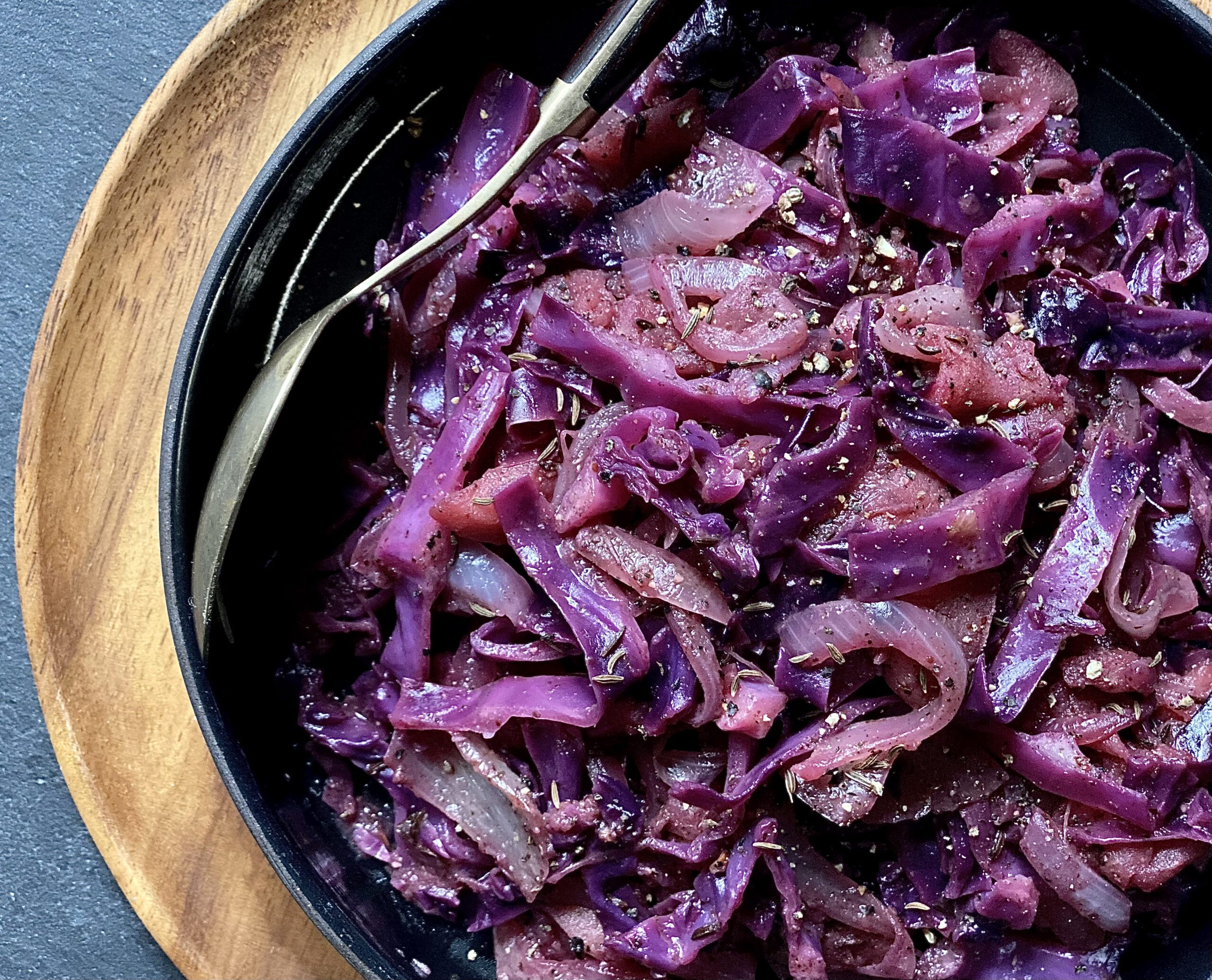 An image showcasing a bowl of braised red cabbage. The cabbage appears tender and slightly wilted, infused with savory spices and aromatics. Specks of seasoning and herbs are visible throughout the dish, adding depth to its appearance.