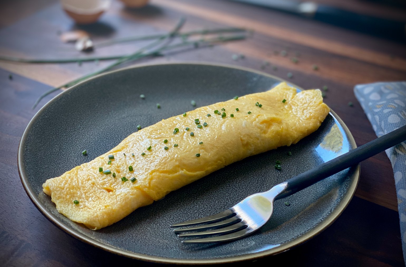 Golden, slightly crisp exterior omelet with a smooth, custardy interior that is topped with chives on a black plate with a fork.