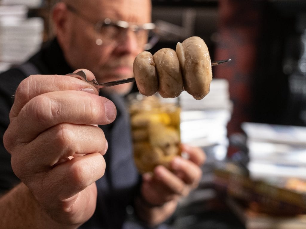 Alton Brown holding three pickled mushrooms on a skewer.