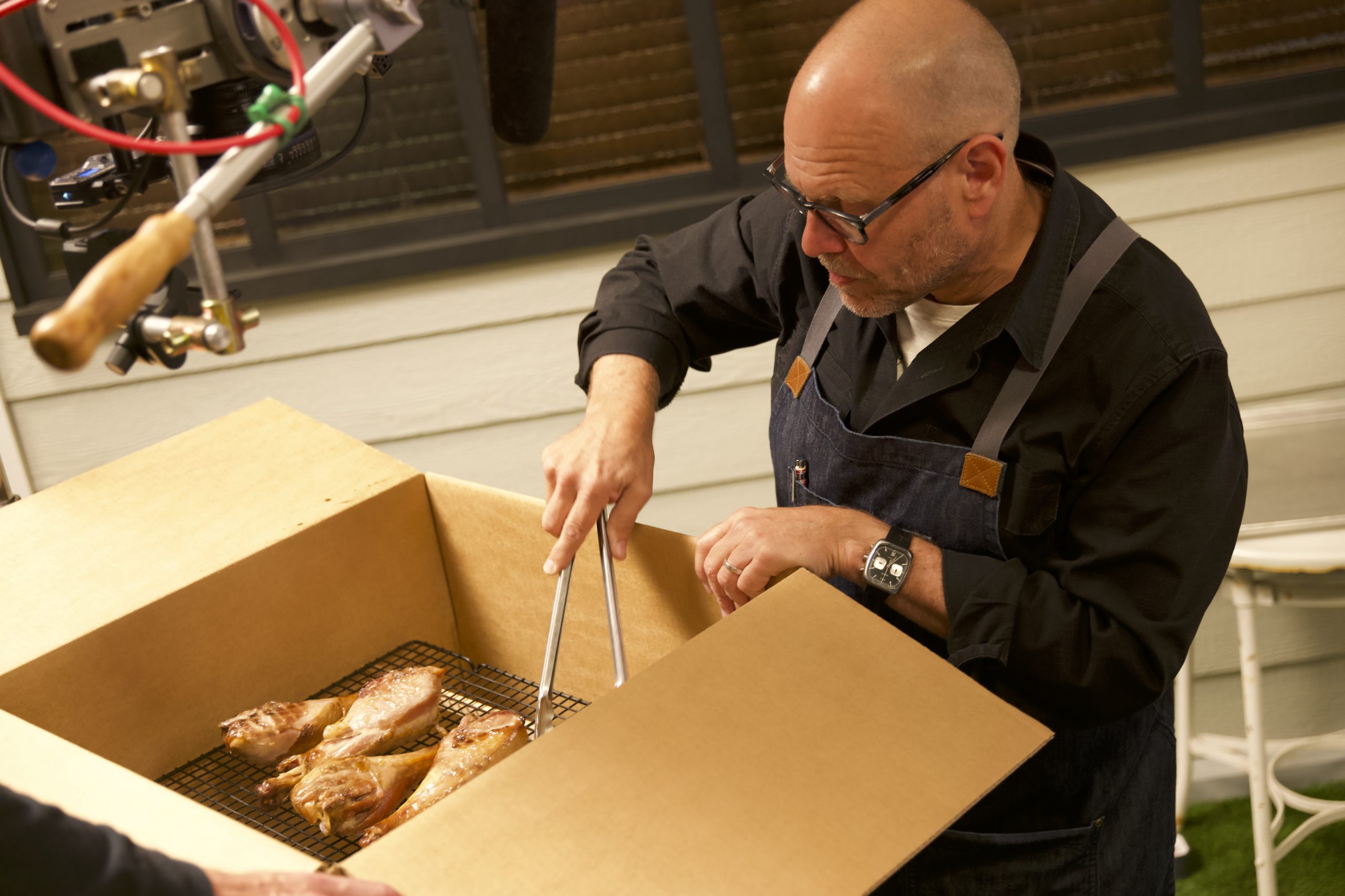 Alton Brown uses tongs to flip Smoked Turkey Legs inside his Cardboard Box Smoker.