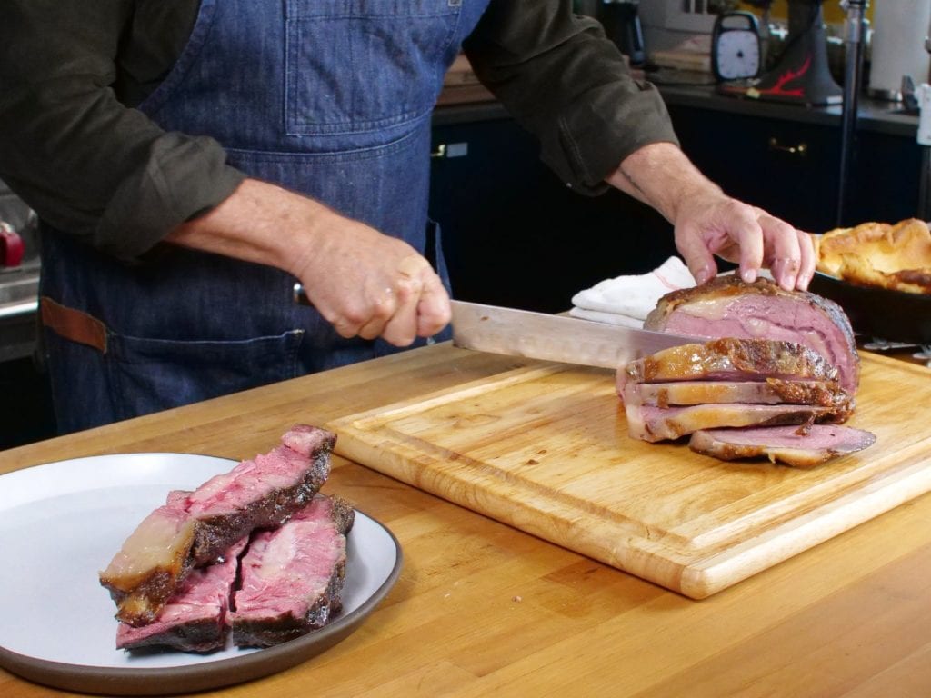 Man wearing apron cutting thick & juicy cuts of rib roast on a wood cutting board.