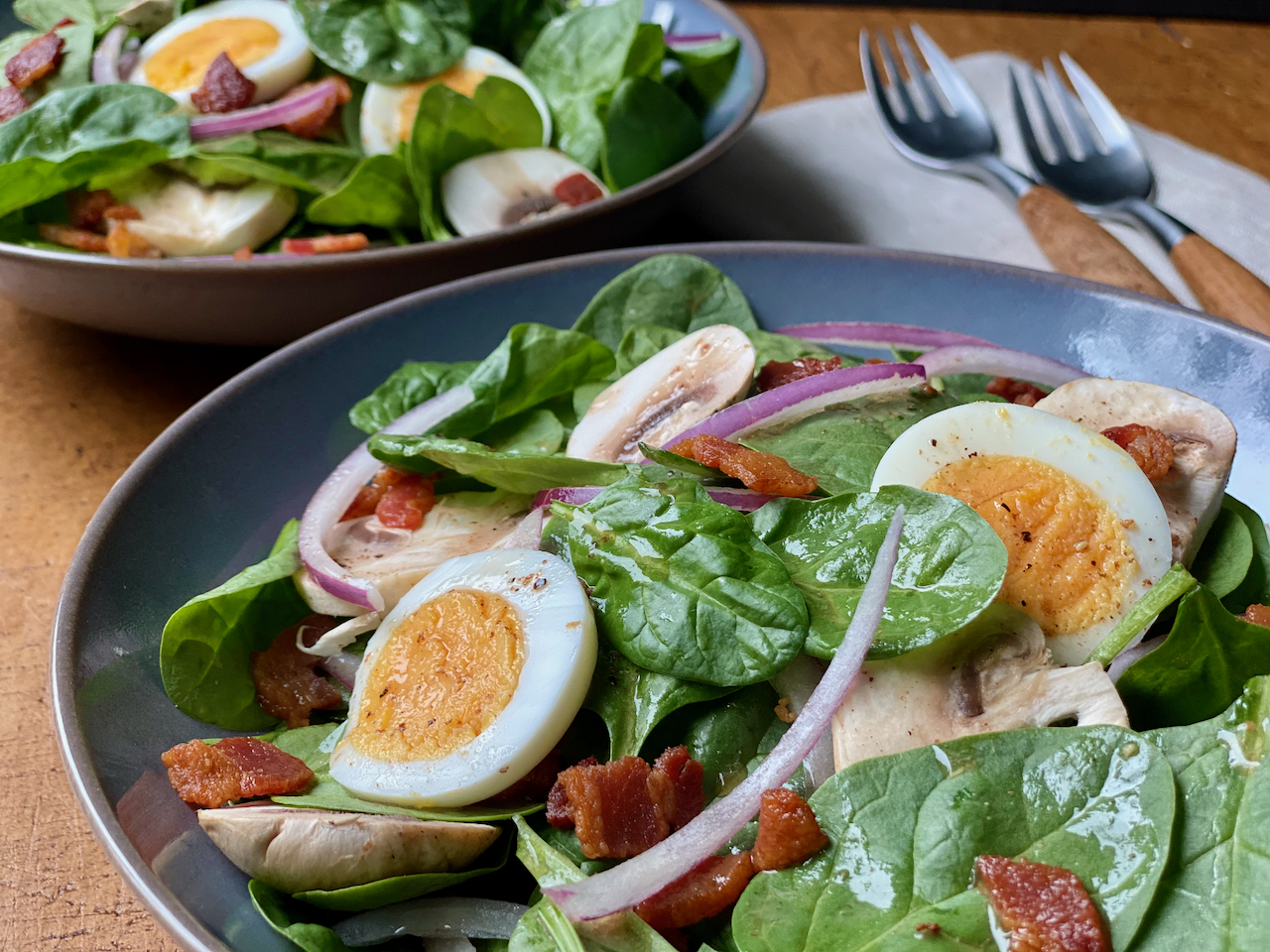 Bowl showing a serving of spinach salad with warm bacon dressing. The salad features vibrant green spinach leaves arranged neatly, topped with crispy bacon, sliced hard boiled eggs and sliced red onions.