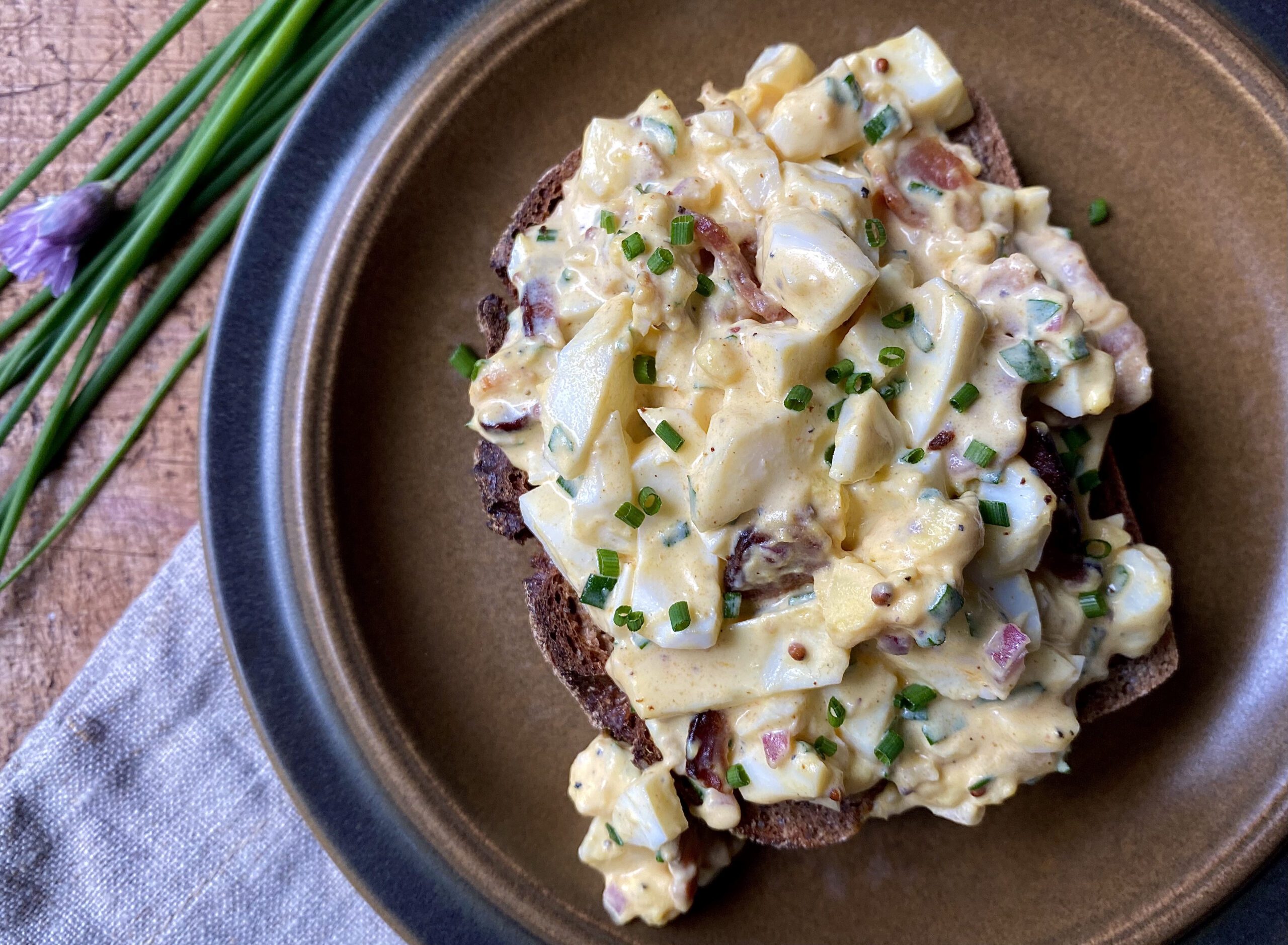 Creamy egg salad with bacon, onion, and parsley that is served on crusty toast and garnished with chives
